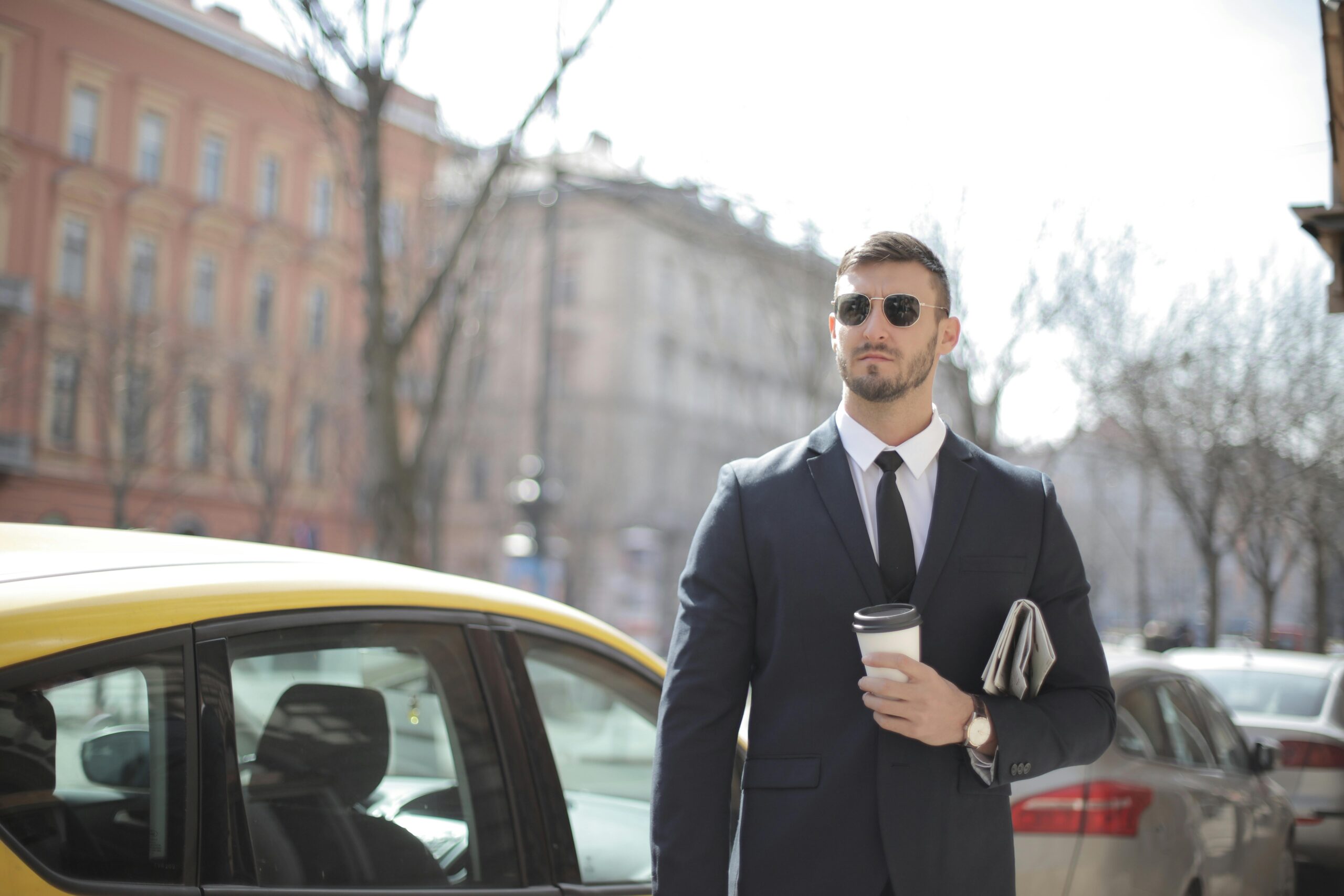 A confident businessman in a suit stands outdoors, holding coffee.